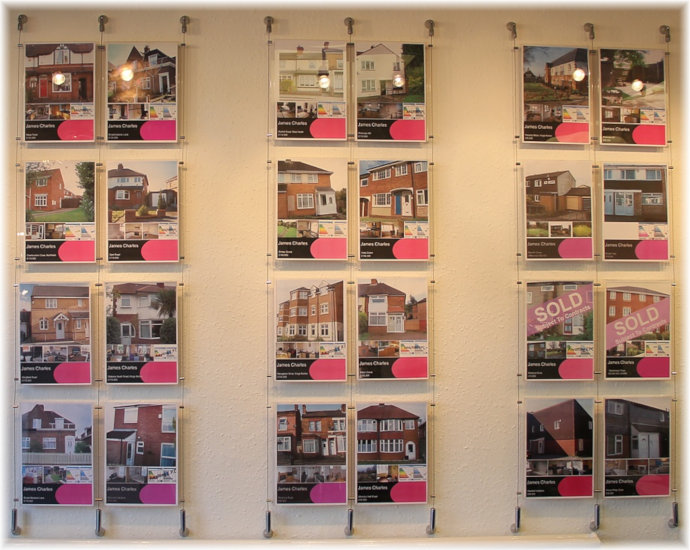 Three wall mounted cable-suspended poster holder displays side-by-side, photographed from inside the estate agent’s reception. Ceiling spotlights are shining onto the displays.